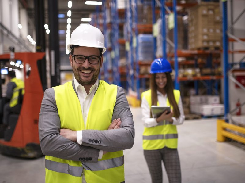 Portrait of successful warehouse worker or supervisor with crossed arms standing in large storage distribution area.